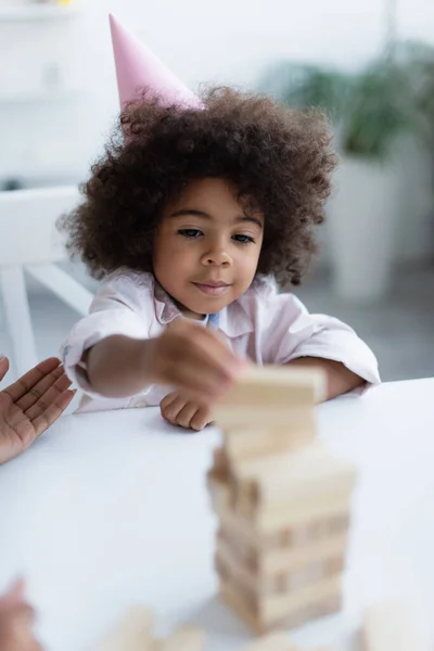 Frisé afro-américain enfant en chapeau de fête jouer jeu blocs de bois sur flou premier plan — Photo de stock