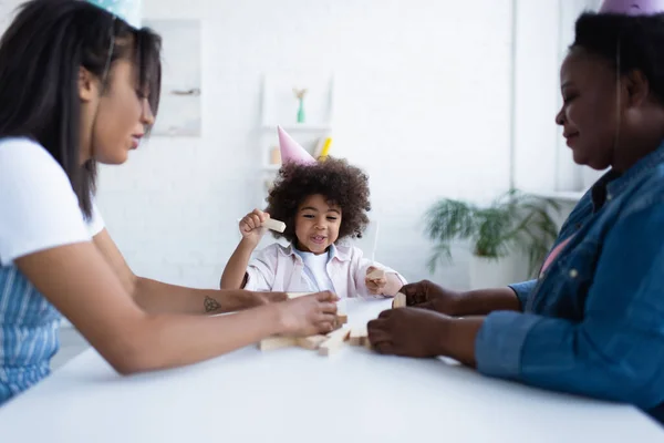 Positivo africano americano chica en partido gorra jugando madera bloques juego con borrosa mamá y abuelita - foto de stock