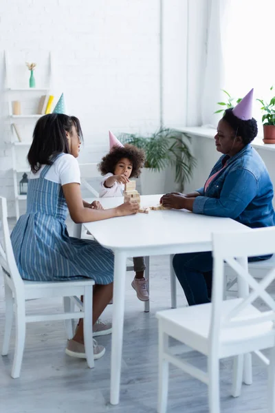 African american girl with mom and granny in party caps playing wood blocks game at home — Stock Photo