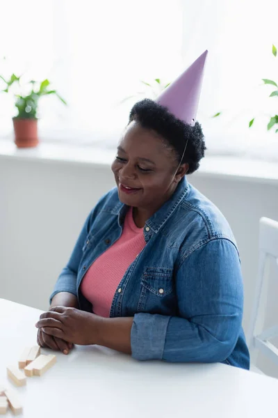 Middle aged african american woman in party cap smiling while sitting near wood blocks game on table — Stock Photo
