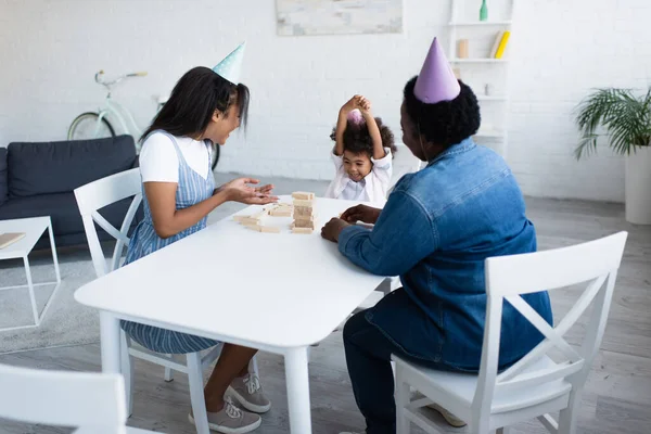 African american woman in party cap pointing at wooden tower on table near happy daughter — Stock Photo