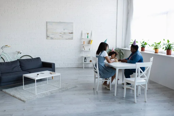 African american women and toddler girl in party caps playing wood blocks game in spacious living room — Stock Photo