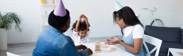 Happy african american girl playing wood blocks game with mother and granny in party caps, banner — Stock Photo