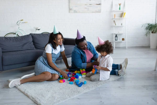 Criança africana americana menina com a mãe e avó em bonés de festa brincando com blocos de construção no chão perto do sofá — Fotografia de Stock