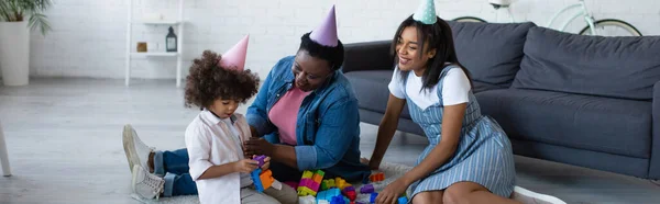 Mujeres afroamericanas felices y niños en gorras de fiesta jugando con bloques de construcción en el piso, pancarta - foto de stock