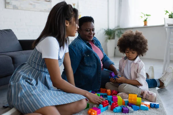 Ricci africano americano ragazza giocare con i blocchi di costruzione vicino madre e nonna al piano a casa — Foto stock