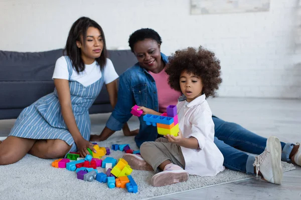 Happy african american women looking at girl playing with building blocks on floor carpet — Stock Photo