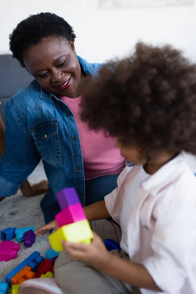 Smiling african american granny looking at blurred granddaughter playing with building blocks — Stock Photo