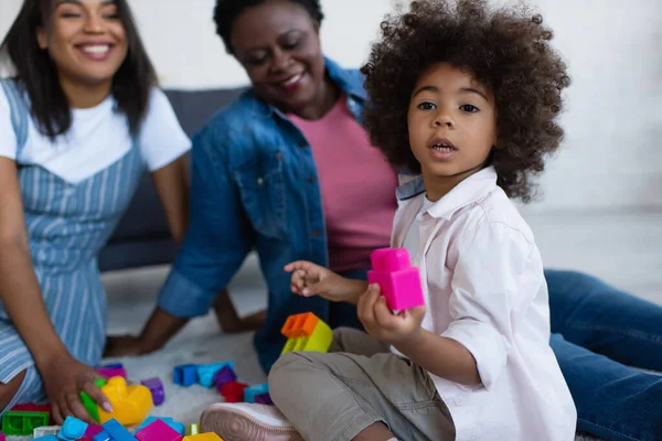 Encaracolado Africano americano menina segurando bloco de construção perto de mãe e avó sorrindo no fundo borrado — Fotografia de Stock