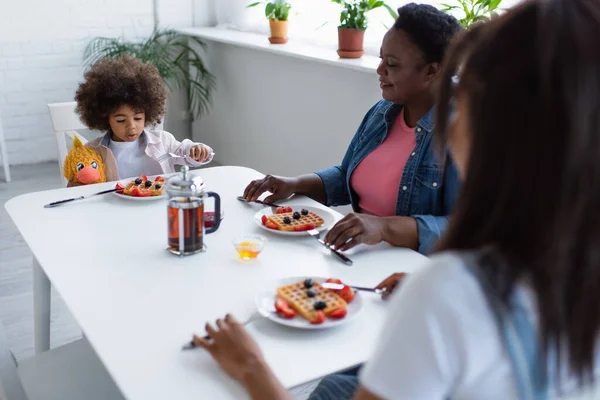 Menina americana africana tomando café da manhã com vovó e mãe borrada enquanto sentado à mesa com brinquedo macio — Fotografia de Stock