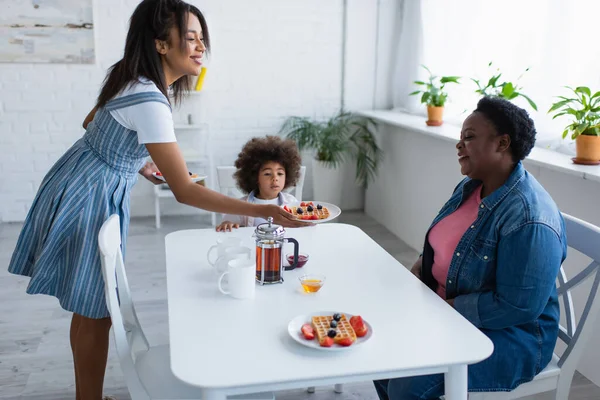 Sonriente afroamericana mujer sosteniendo plato con gofre y bayas cerca de mamá y su hija - foto de stock