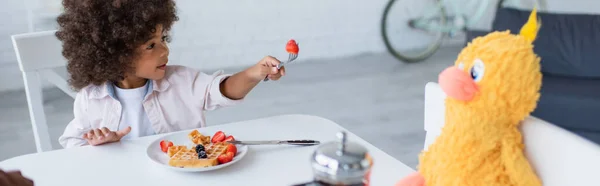 Africano americano niño proponiendo fresa a juguete polluelo durante el desayuno en cocina, pancarta - foto de stock