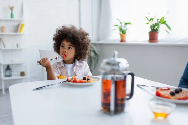 Africano americano ragazza avendo colazione vicino offuscata teiera su tavolo da cucina — Foto stock