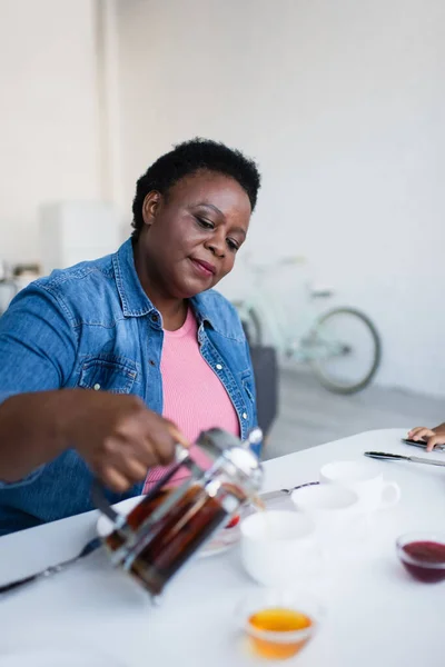 Reife Afroamerikanerin gießt Tee in der Nähe verschwommener Schüsseln mit Marmelade und Honig auf den Tisch — Stockfoto