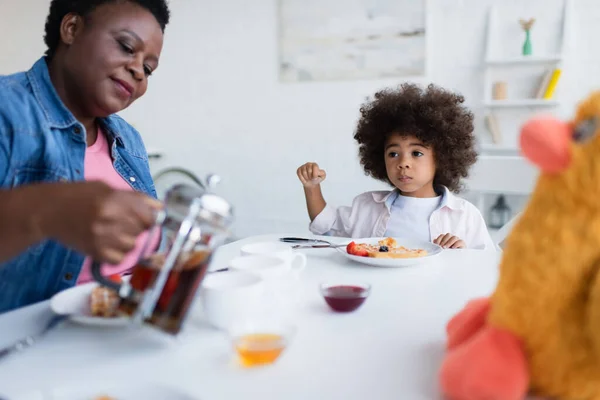 Lockiges afrikanisches Mädchen frühstückt in der Nähe von verschwommenem Stofftier und Oma gießt Tee — Stockfoto