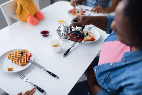 Aus der Vogelperspektive: verschwommene afrikanisch-amerikanische Frau gießt beim Frühstück mit der Familie Tee in der Nähe von leckeren Waffeln mit Beeren — Stockfoto