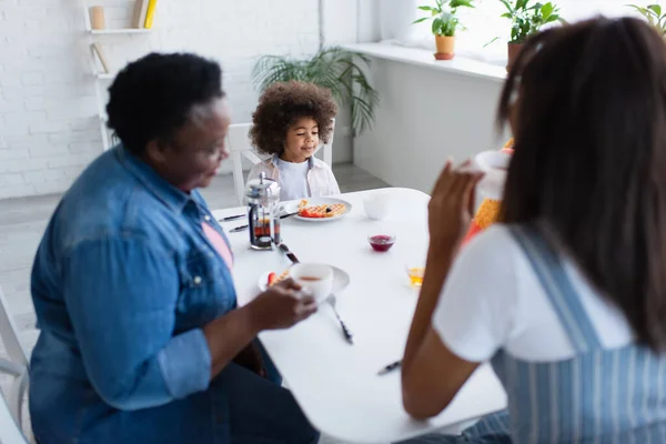 Verschwommene afrikanisch-amerikanische Frauen trinken Tee während des Frühstücks mit Kleinkind Mädchen in der Küche — Stockfoto