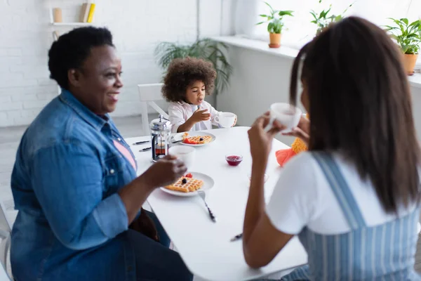 Toddler african american girl having breakfast with smiling granny and blurred mom in kitchen — Stock Photo