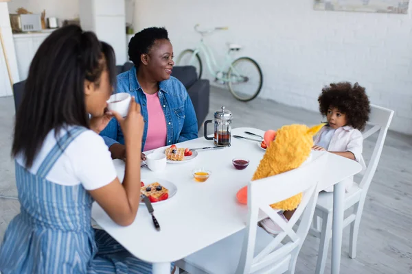 Menina americana africana tomando café da manhã perto de mãe, avó e brinquedo macio na cozinha — Fotografia de Stock