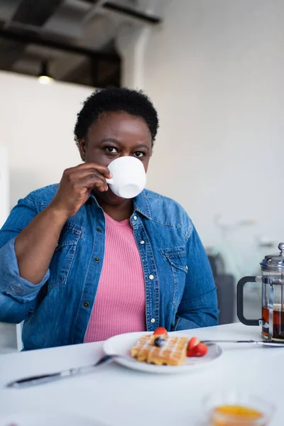 Afrikanische Amerikanerin mittleren Alters trinkt Tee in der Nähe einer verschwommenen Waffel mit frischen Beeren auf dem Teller — Stock Photo