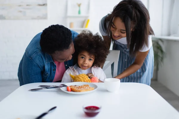 Feliz africano americano mujeres abrazando niño sentado con suave juguete cerca de waffle y bowl con mermelada - foto de stock