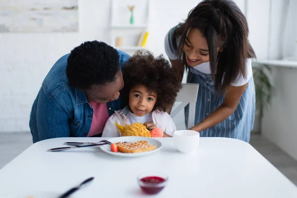 Mulheres americanas africanas felizes abraçando criança sentada com brinquedo macio perto de waffle e tigela com geléia — Fotografia de Stock