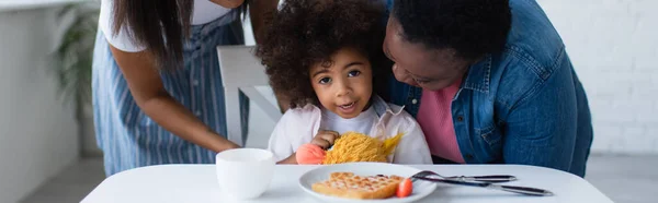 Mujeres afroamericanas abrazando a un niño sentado con un juguete suave cerca de la placa con gofre en la mesa, pancarta - foto de stock