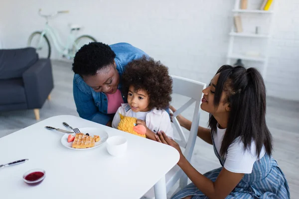Mujeres afroamericanas felices abrazando al niño sentado cerca del gofre con bayas y mermelada en la mesa - foto de stock