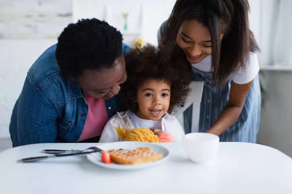 Alegre Africano americano menina sentada à mesa com café da manhã borrado perto sorrindo vovó e mãe — Fotografia de Stock