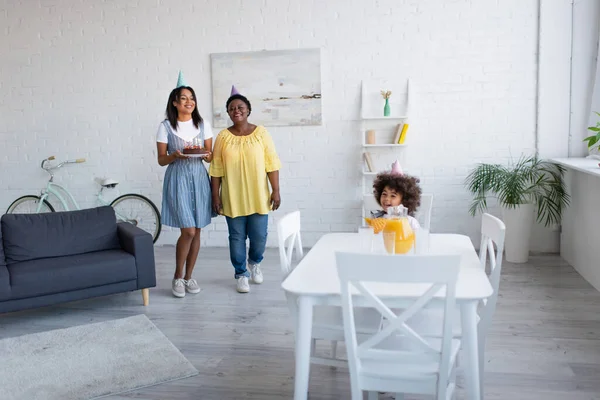 Femmes afro-américaines avec gâteau d'anniversaire près fille heureuse dans le chapeau de fête assis à table avec cruche de jus d'orange frais — Photo de stock