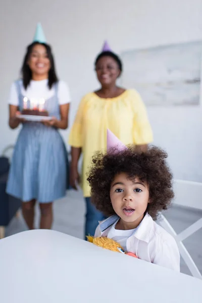 Rizado africano americano chica mirando cámara cerca borrosa mamá y abuelita con pastel de cumpleaños - foto de stock