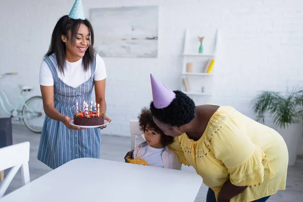 Africano americano ragazza seduta a tavolo vicino nonna abbracciare lei e madre tenendo compleanno torta — Foto stock