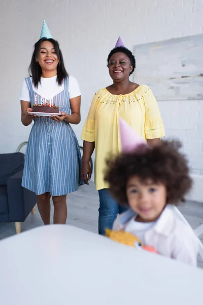 Borrosa africana americana chica sentado en sala de estar cerca de mamá y la abuela con pastel de cumpleaños - foto de stock