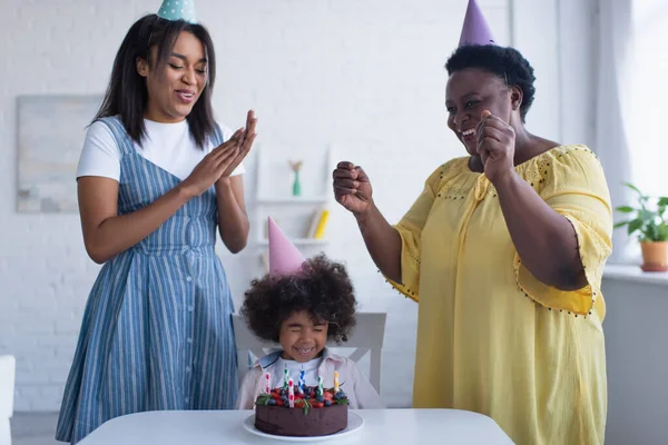 Niño afroamericano chica con los ojos cerrados sentado cerca de pastel de cumpleaños y mamá con la abuela en gorras de fiesta — Stock Photo