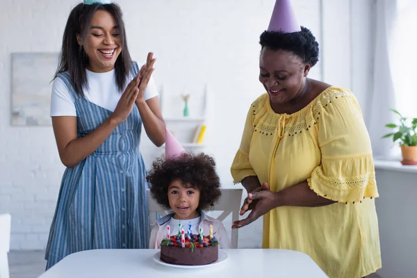 Alegres mujeres afroamericanas aplaudiendo de la mano cerca de niña y pastel de cumpleaños — Stock Photo