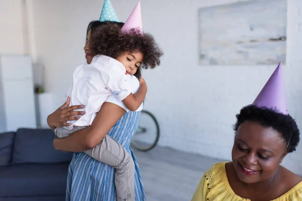 African american mother and daughter in party caps embracing near smiling granny — Stock Photo