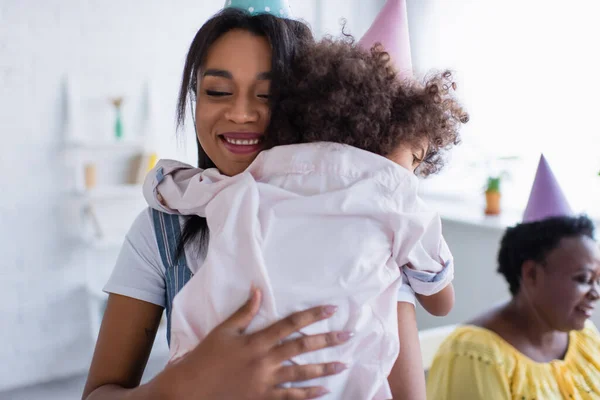 Glücklich afrikanisch-amerikanische Frau mit geschlossenen Augen umarmt Kleinkind Tochter in der Nähe verschwommene Oma in Party-Mütze — Stockfoto