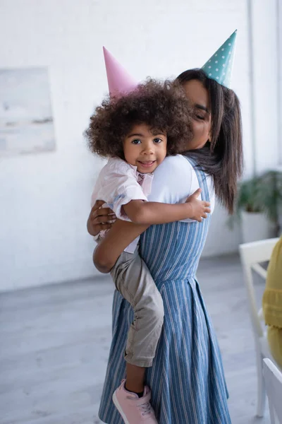 Jeune mère afro-américaine et fille heureuse dans les casquettes de fête embrassant à la maison — Photo de stock