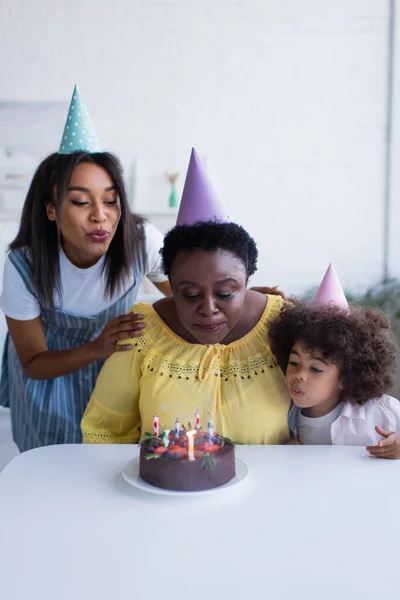 Mujeres afroamericanas y niña en gorras de fiesta soplando velas en pastel de cumpleaños - foto de stock