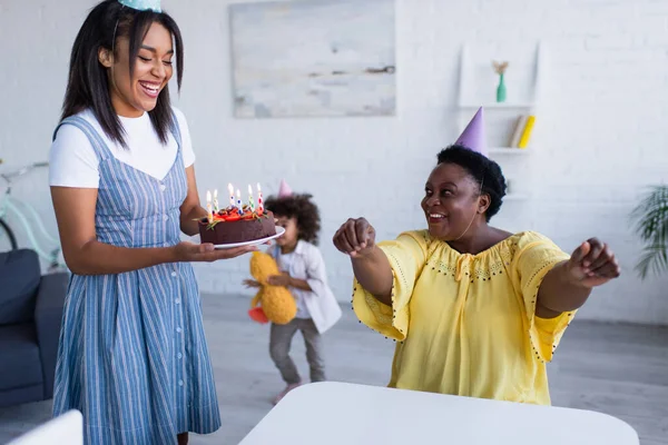 Jovem afro-americana segurando bolo de aniversário perto de mãe feliz em boné de festa — Fotografia de Stock