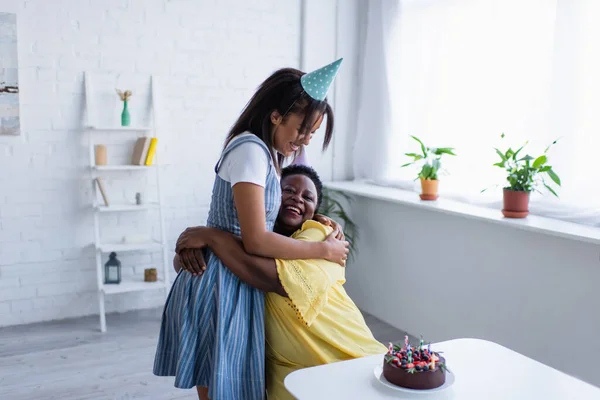 Giovane donna afroamericana in berretto da festa che abbraccia la madre felice vicino alla torta di compleanno con le candele — Foto stock