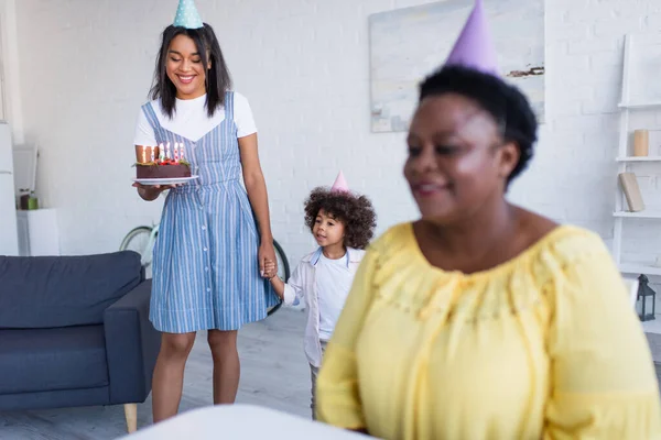 Lächelnde afrikanisch-amerikanische Mutter und Tochter mit Geburtstagstorte neben verschwommener Oma in Partymütze — Stockfoto