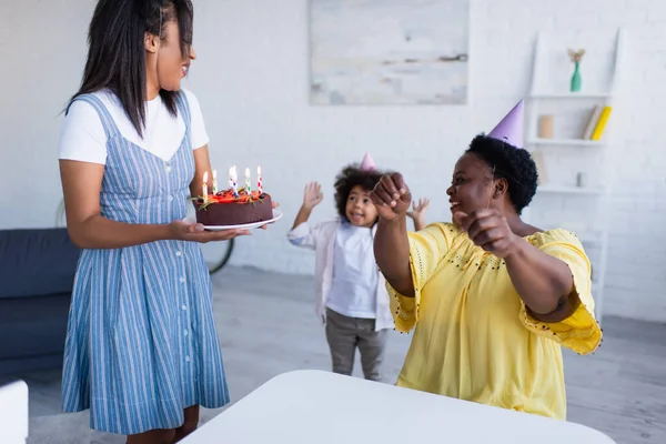 Молода афроамериканка тримає торт на день народження біля веселої дочки і бабусі в партійних шапках — стокове фото