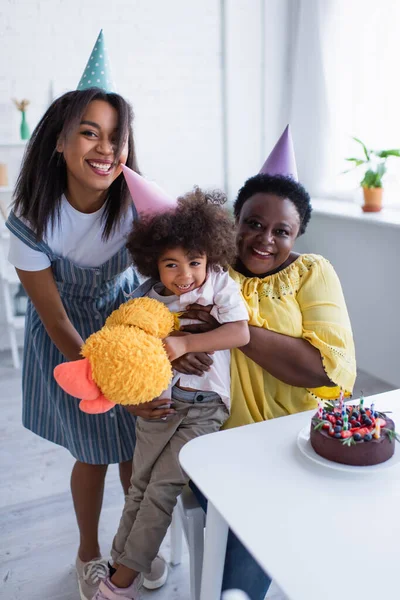 Веселая африканская американка держит мягкую игрушку, в то время как мама и бабушка обнимают ее около праздничного торта — стоковое фото