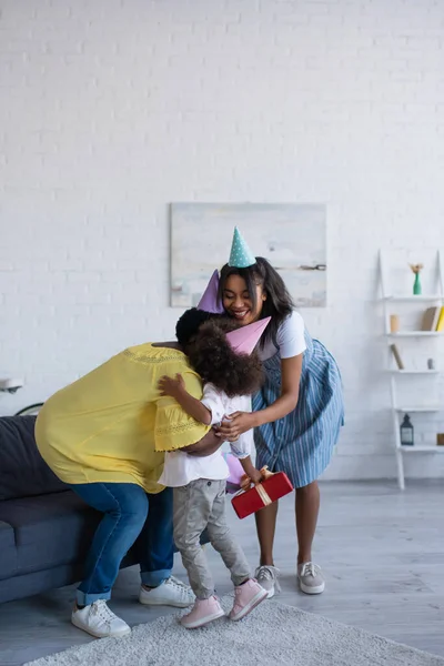 Niña pequeña sosteniendo regalo de cumpleaños mientras la abuela y la mamá en gorras de fiesta abrazándola en la sala de estar - foto de stock