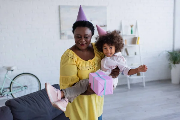 Maduro africano americano mujer en partido gorra celebración alegre nieta y regalo caja en manos - foto de stock