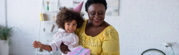 Joyeuse femme afro-américaine d'âge moyen tenant sa petite-fille dans une casquette de fête et une boîte-cadeau, une bannière — Photo de stock