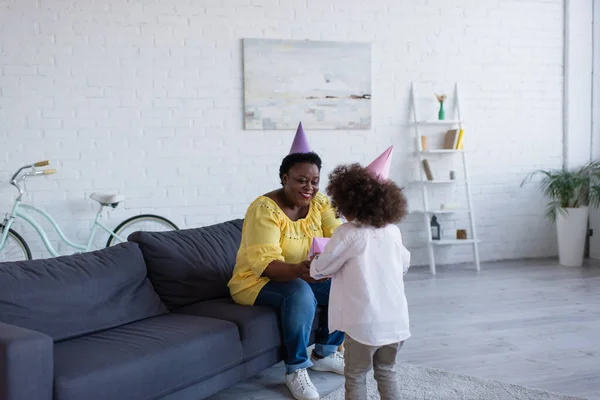 Rückseite des lockigen Mädchens mit Partymütze präsentiert Geschenk an afrikanisch-amerikanische Oma auf dem Sofa im Wohnzimmer sitzen — Stockfoto