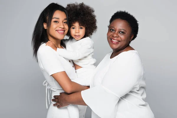 Pleased african american women in white clothes looking at camera while holding child isolated on grey — Stock Photo