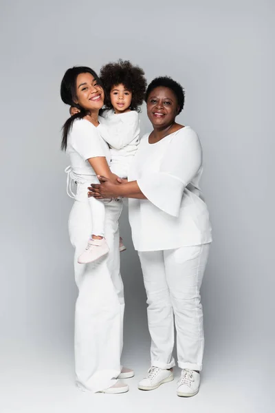 Cheerful african american women in white clothes holding toddler girl on grey — Stock Photo